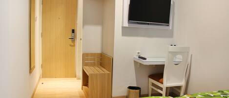 A small, well-lit room with a wooden door, a bed with a leaf-patterned bedspread, a white desk with a chair, and a mounted television.