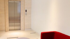 A modern lobby with a red sofa, two round ottomans, a wooden floor, and a wall-mounted elevator. - Stori Hotel Ambon (Ambon)