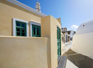 A narrow street with buildings featuring white walls and green doors, leading to a church with a bell tower. - Domenica Home & Spa (Santorini)