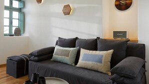 Una sala moderna con un sofá gris oscuro, una mesa de centro redonda y piso de madera. Se observan apliques hexagonales en la pared y un reloj.