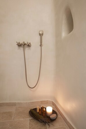 A bathroom with a shower, a stone-tiled floor, and a tray with toiletries and a candle. - Domenica Home & Spa (Santorini)