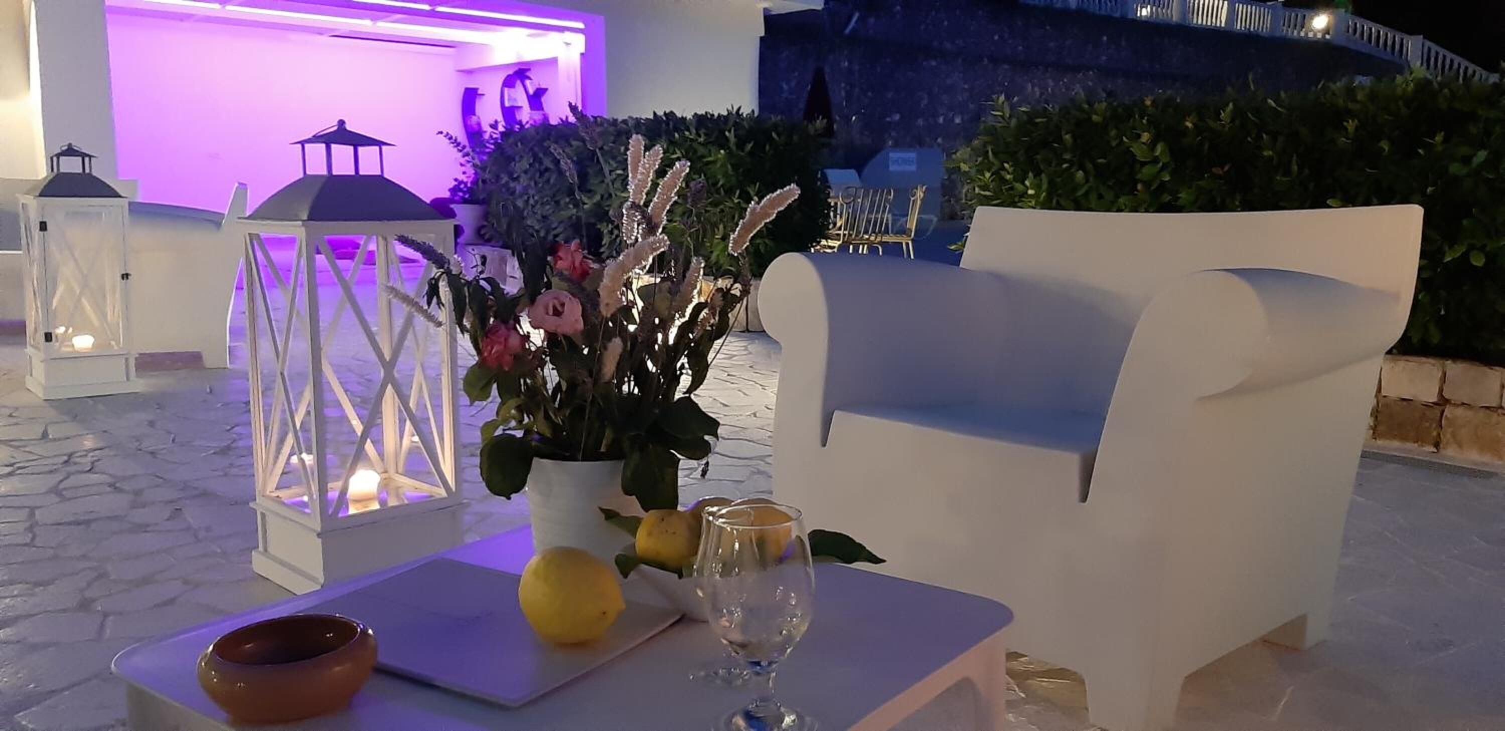 Area salotto esterna con sedie bianche, un tavolo con una ciotola, limoni e bicchieri da vino.