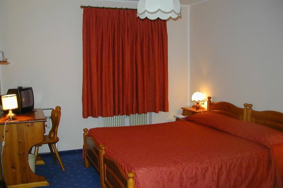 A bedroom with a wooden bed, a desk with a lamp, a chair, and a window with red curtains.