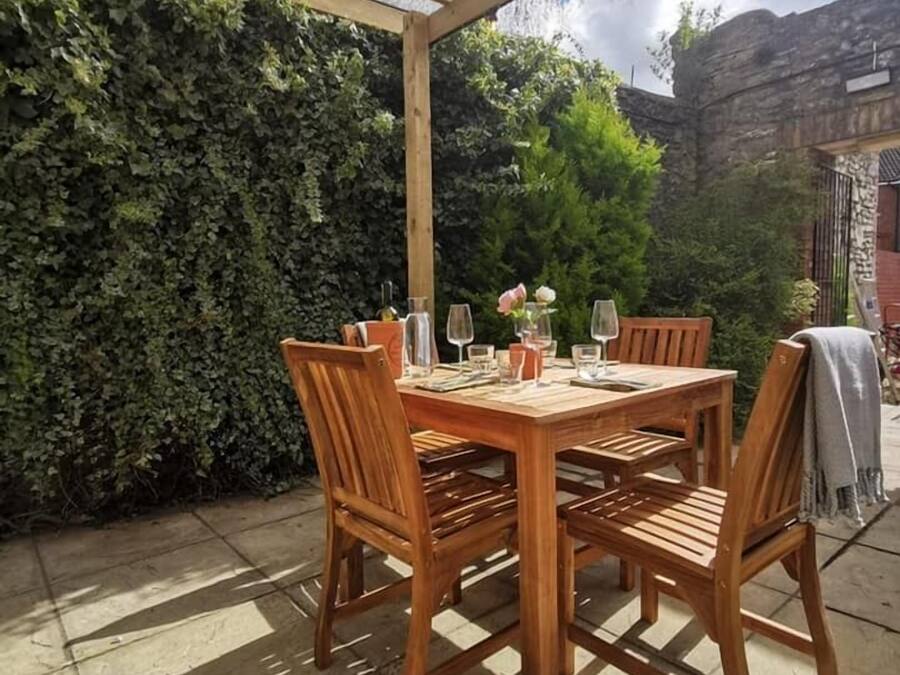 Un juego de mesa y sillas de madera dispuesto bajo una pérgola con techo de vidrio, rodeado de un muro de piedra y abundante vegetación.