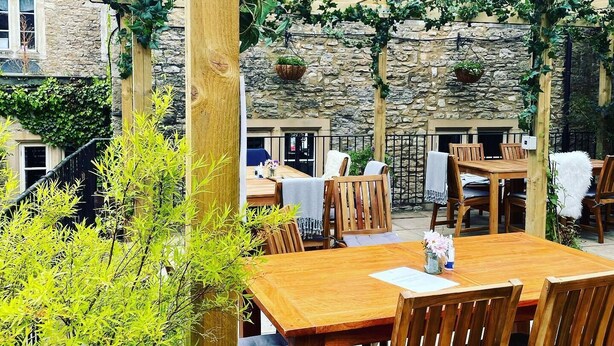 An outdoor patio with wooden tables and chairs, stone walls, and greenery.