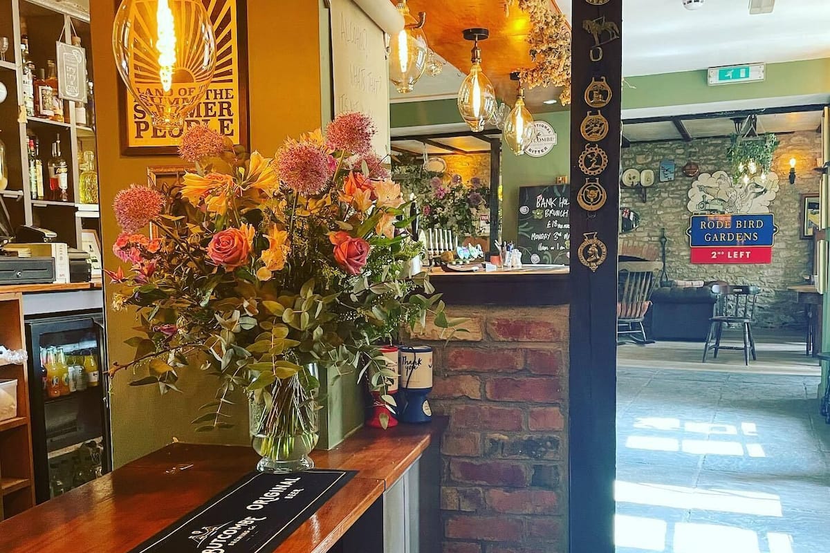 A bar area with a wooden counter, a vase of flowers, hanging light fixtures, and a sign for 'Rode Bird Gardens'.