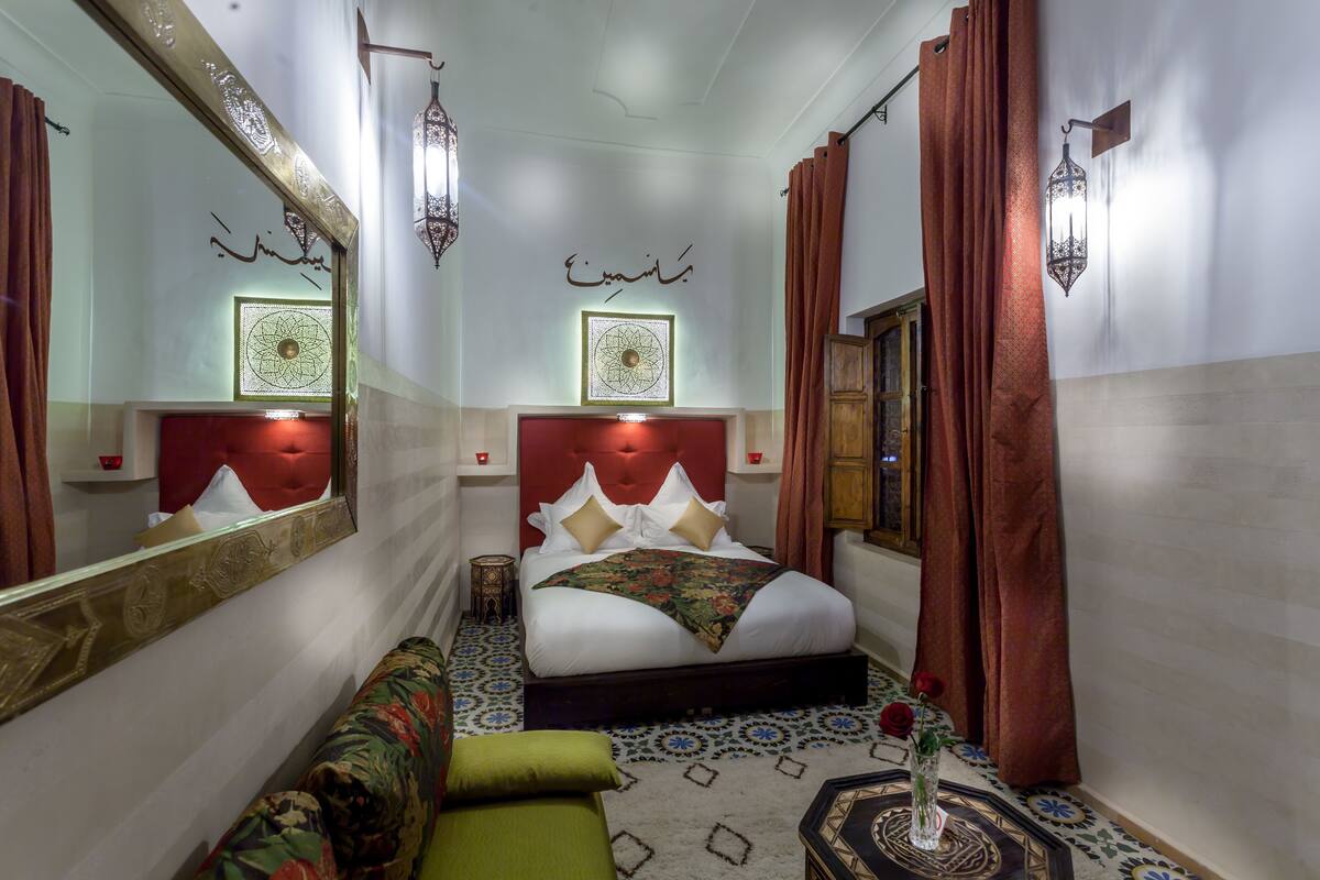 A hotel room with a large bed, a sofa, a decorative mirror, a wall-mounted lamp, and a patterned carpet.