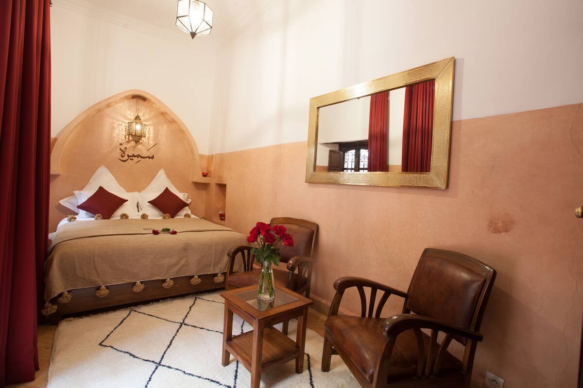 A bedroom with a bed, a chair, a small table, a mirror, and a vase with flowers.
