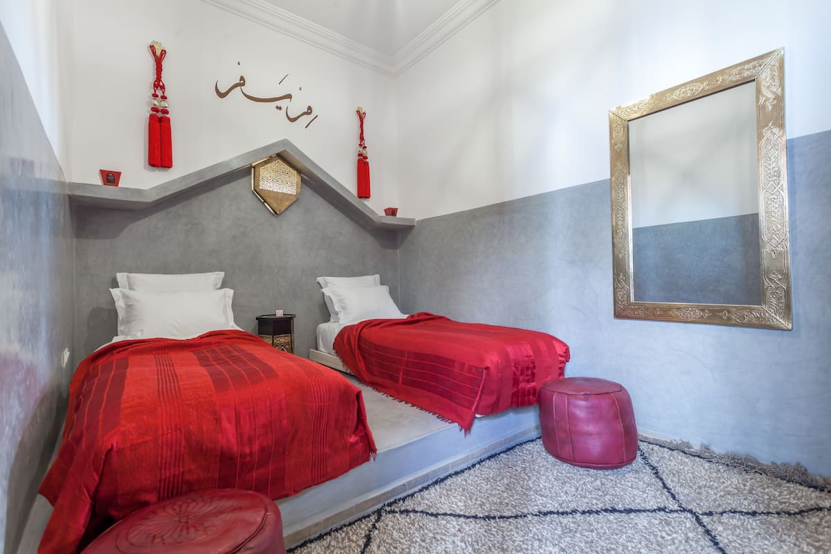 A bedroom with two beds, a red ottoman, and a decorative mirror.