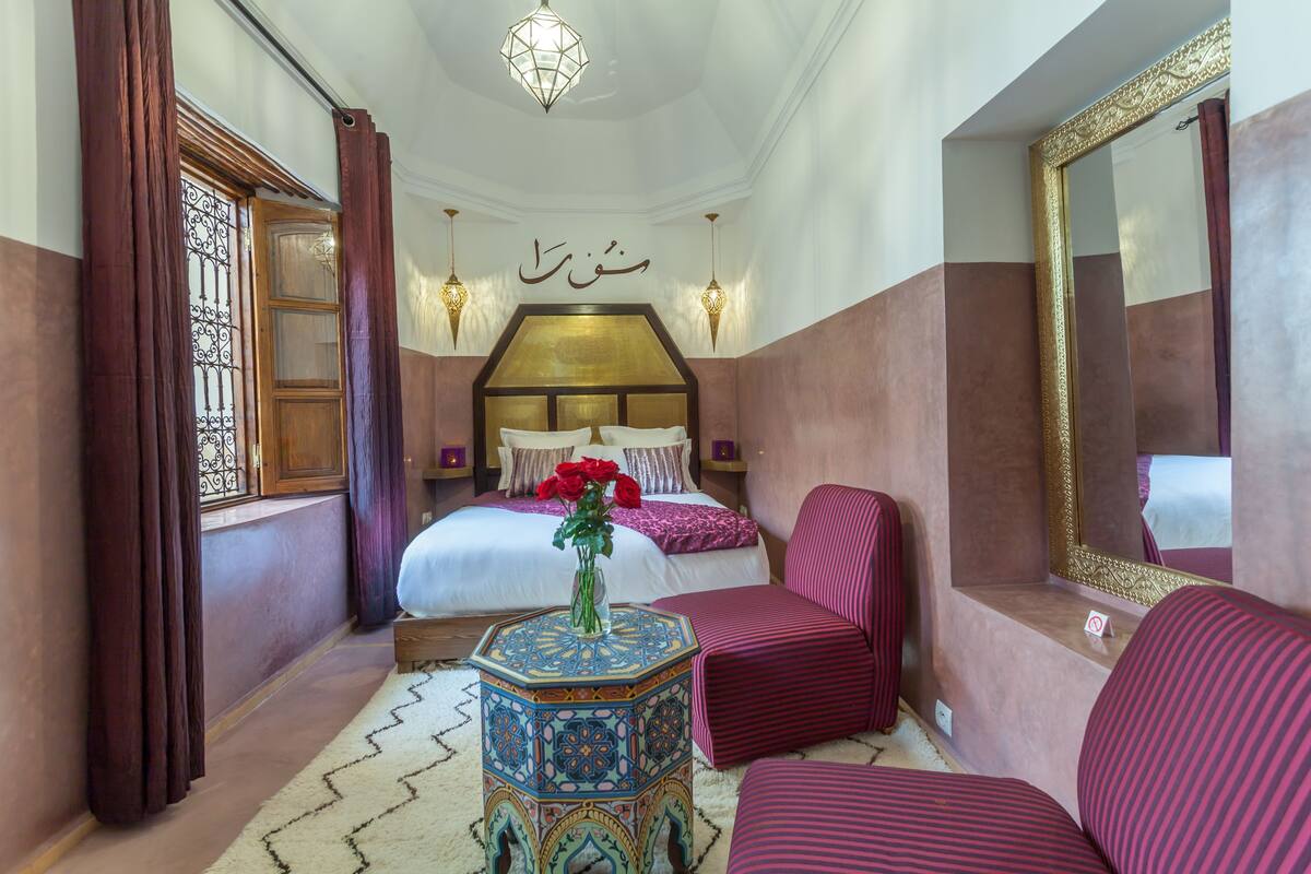 A bedroom with a bed, a bedside table with a vase, a chair, a mirror, and a decorative wall piece.