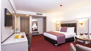 A hotel room with a bed, bedside table, and a desk with fruits and water bottles. - Perla Arya Hotel (Izmir)