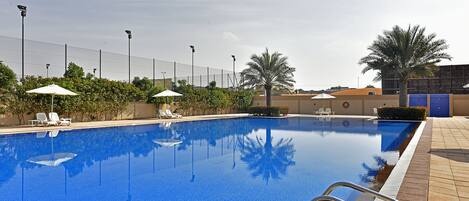 A swimming pool with clear blue water, surrounded by lounge chairs and umbrellas, and a fence in the background.