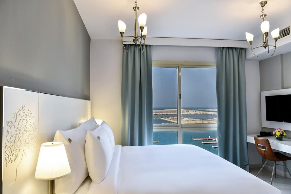 A hotel room with a bed, a desk, a TV, and a view of the sea.