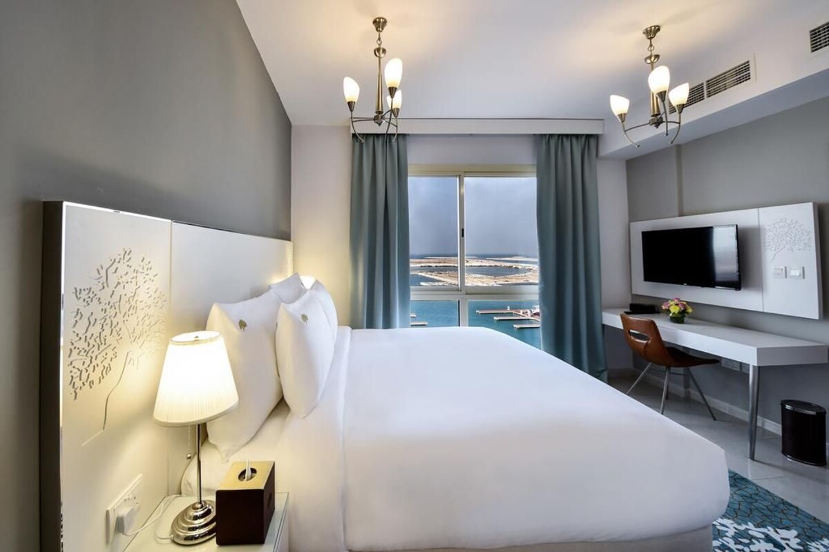 A hotel room with a bed, a desk, a TV, and a view of the sea.