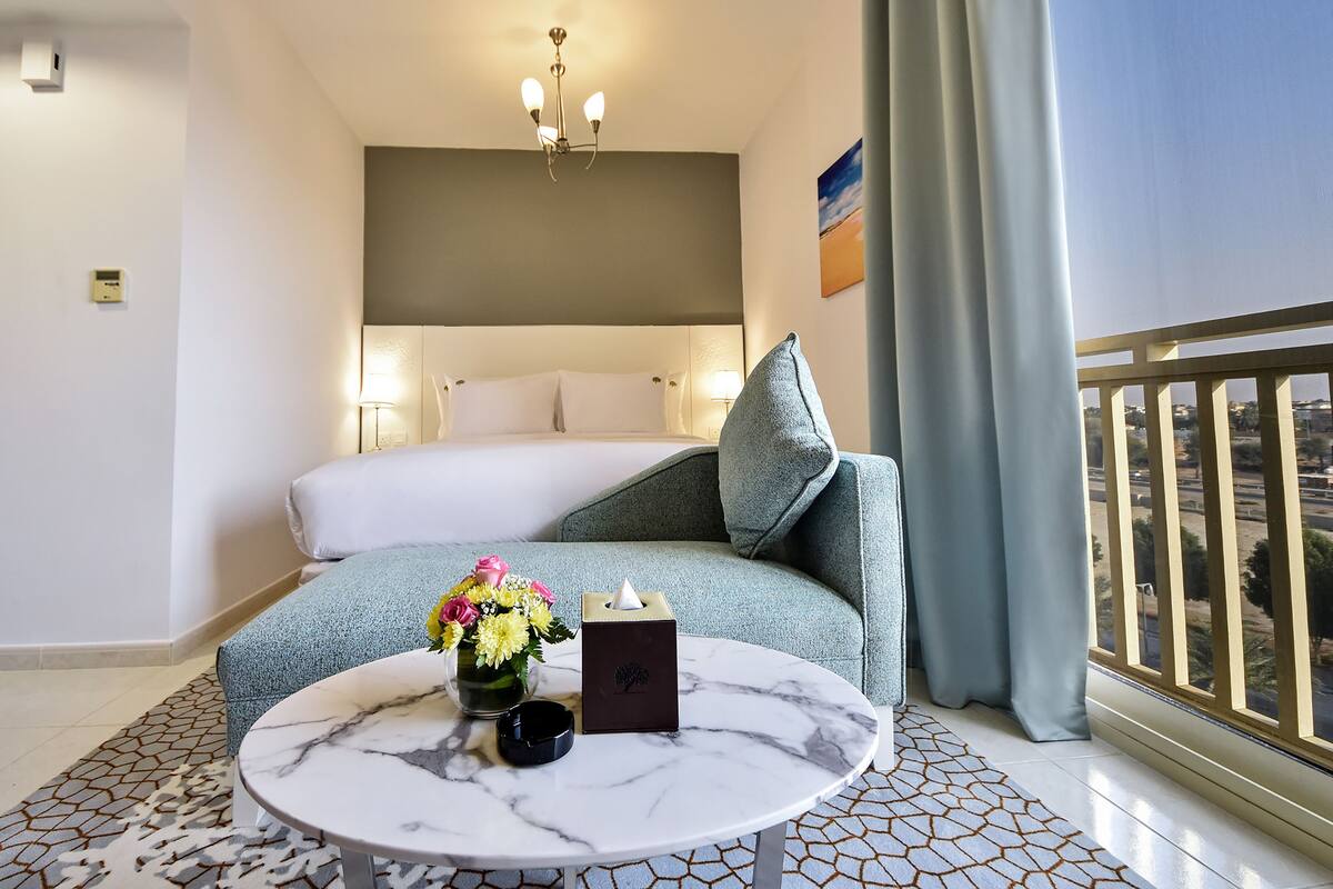 Hotellihuone, jossa on sänky, pyöreä pöytä maljakko täynnä kukkia, yöpöytälamppu ja parvekkeelta avautuva näkymä kaupunkiin.