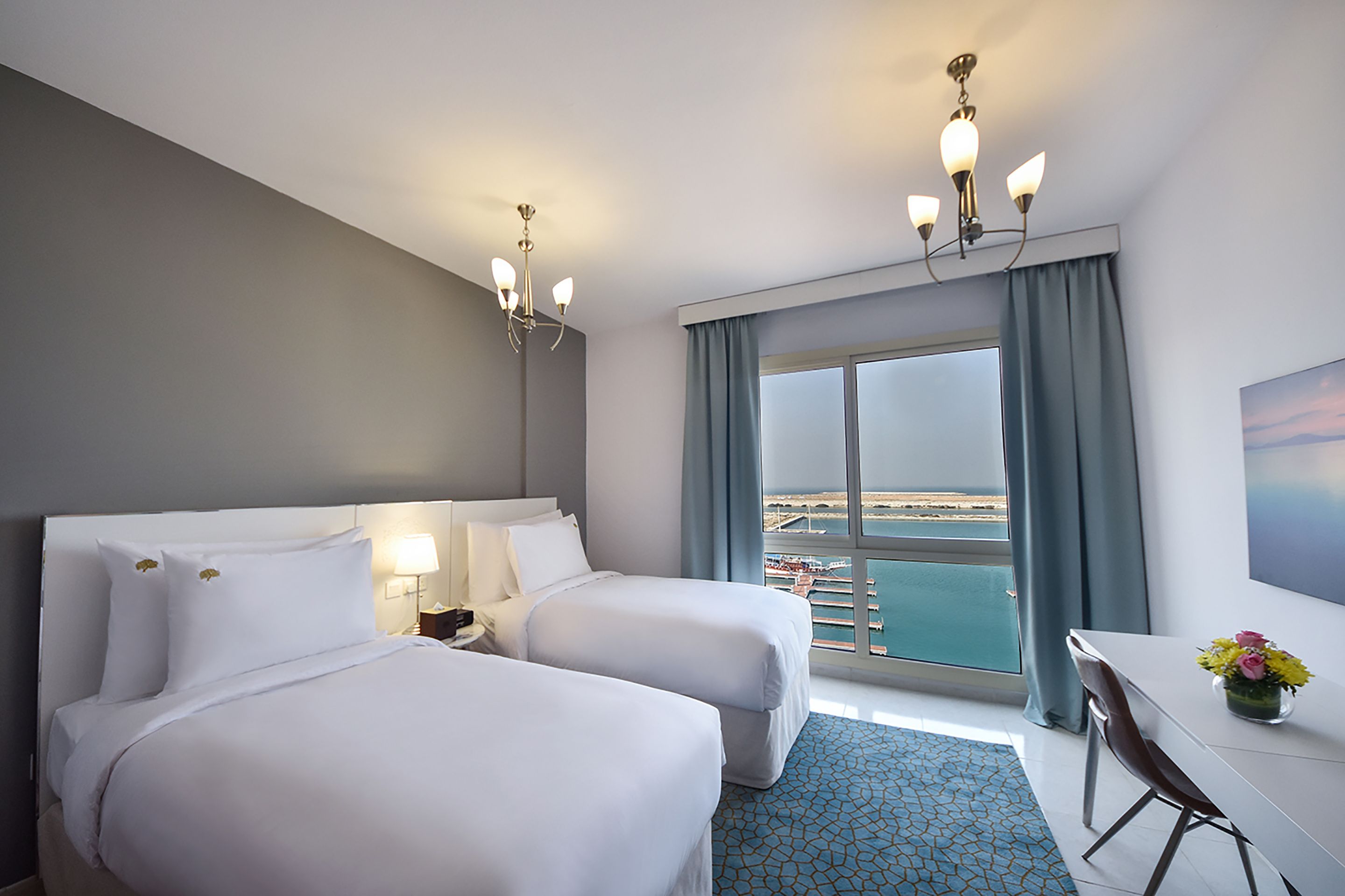 A hotel room with two beds, a desk, and a view of the sea.