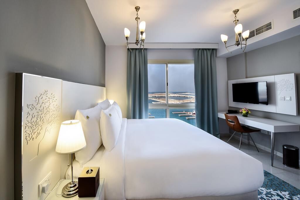 A hotel room with a bed, a desk, a TV, and a view of the sea.