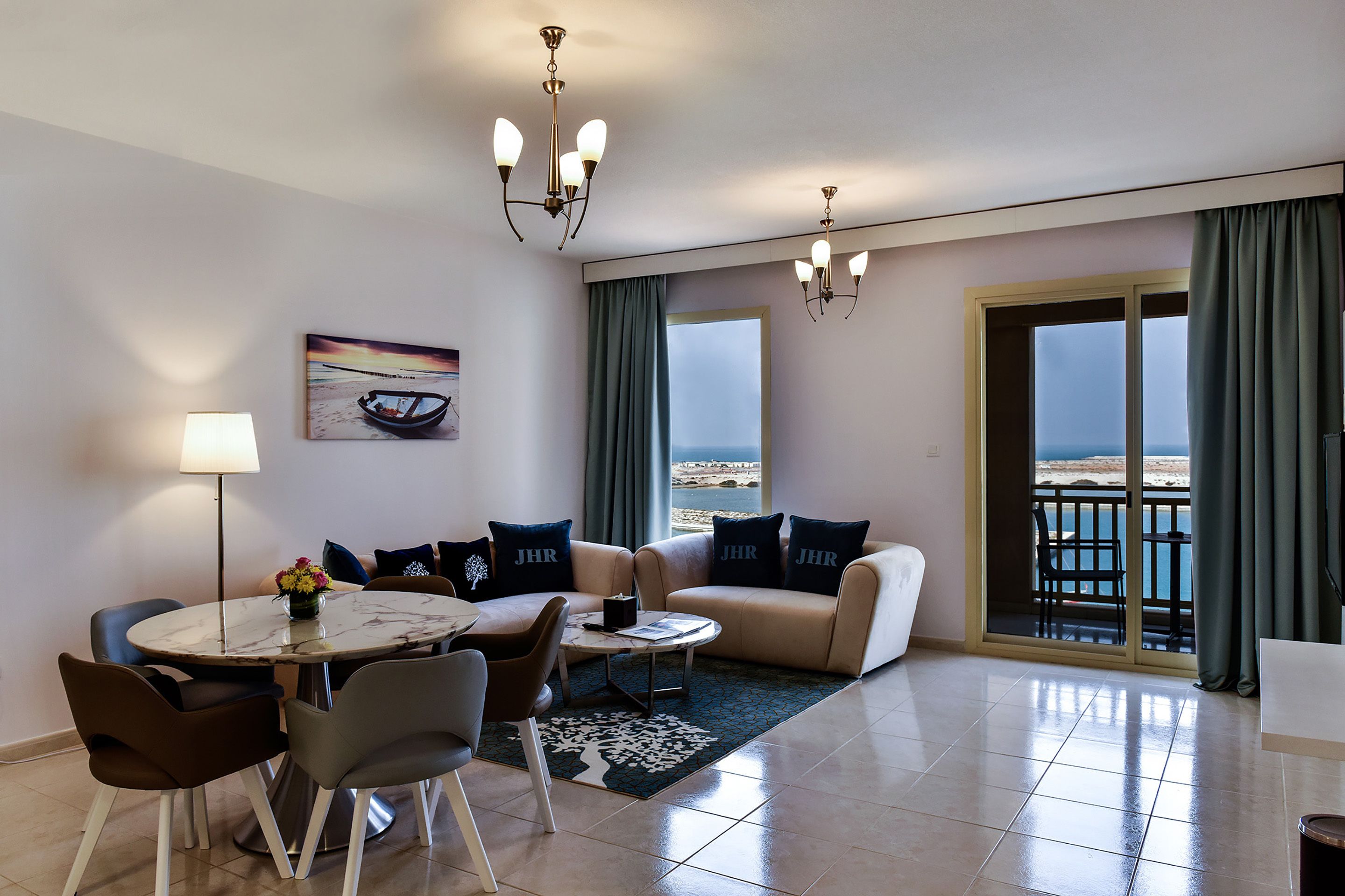 Una sala moderna con comedor, un conjunto de sofá, una araña, un cuadro y vista al mar a través de un ventanal grande.