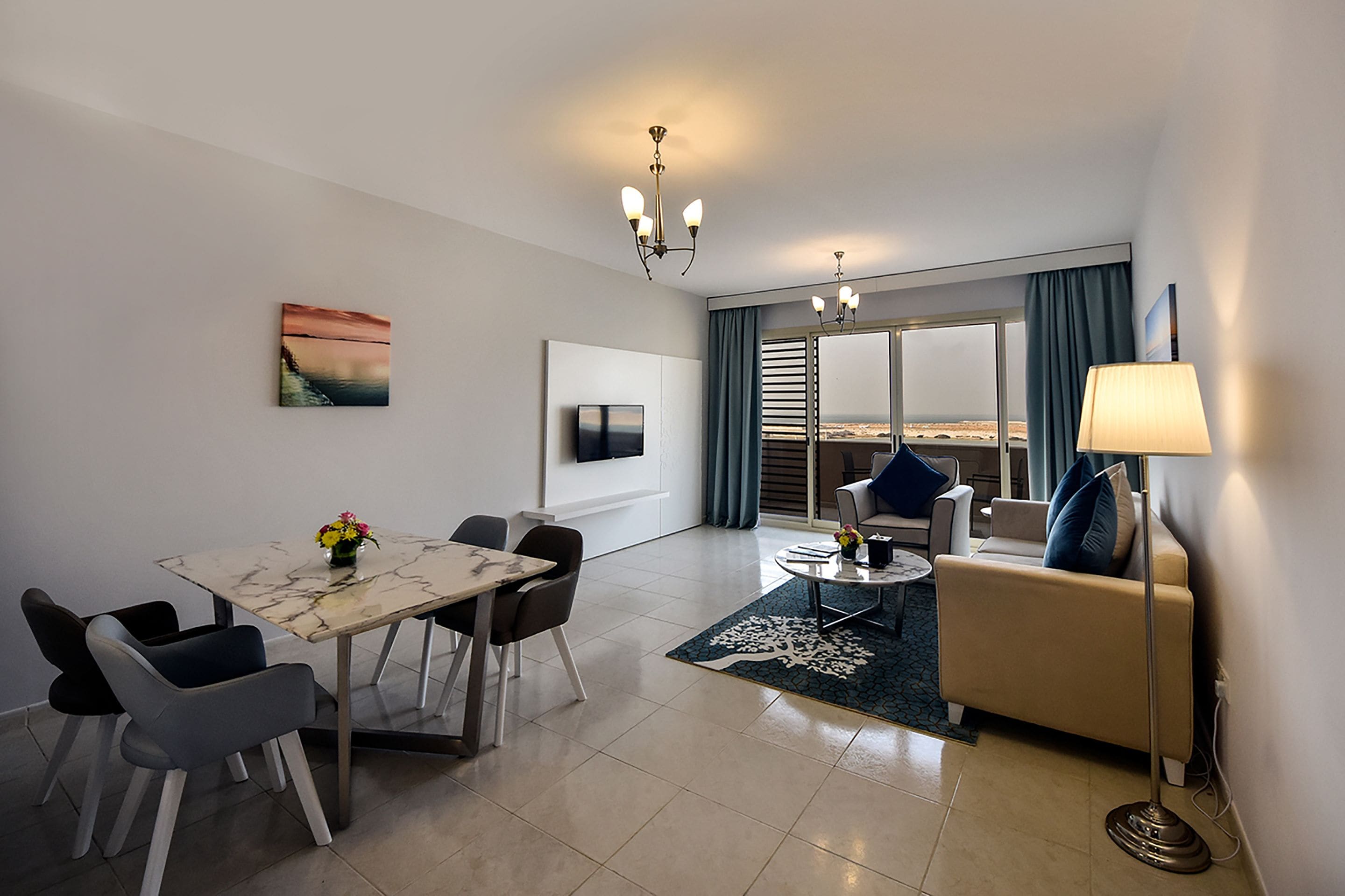 A modern living room with a sofa, a dining table with chairs, a TV mounted on the wall, and a large window with curtains.