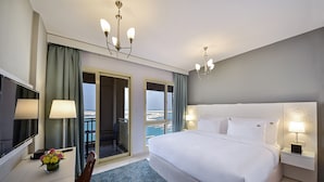 Habitación de hotel con una cama grande, televisión, escritorio con lámpara y vista al mar a través de las puertas del balcón.