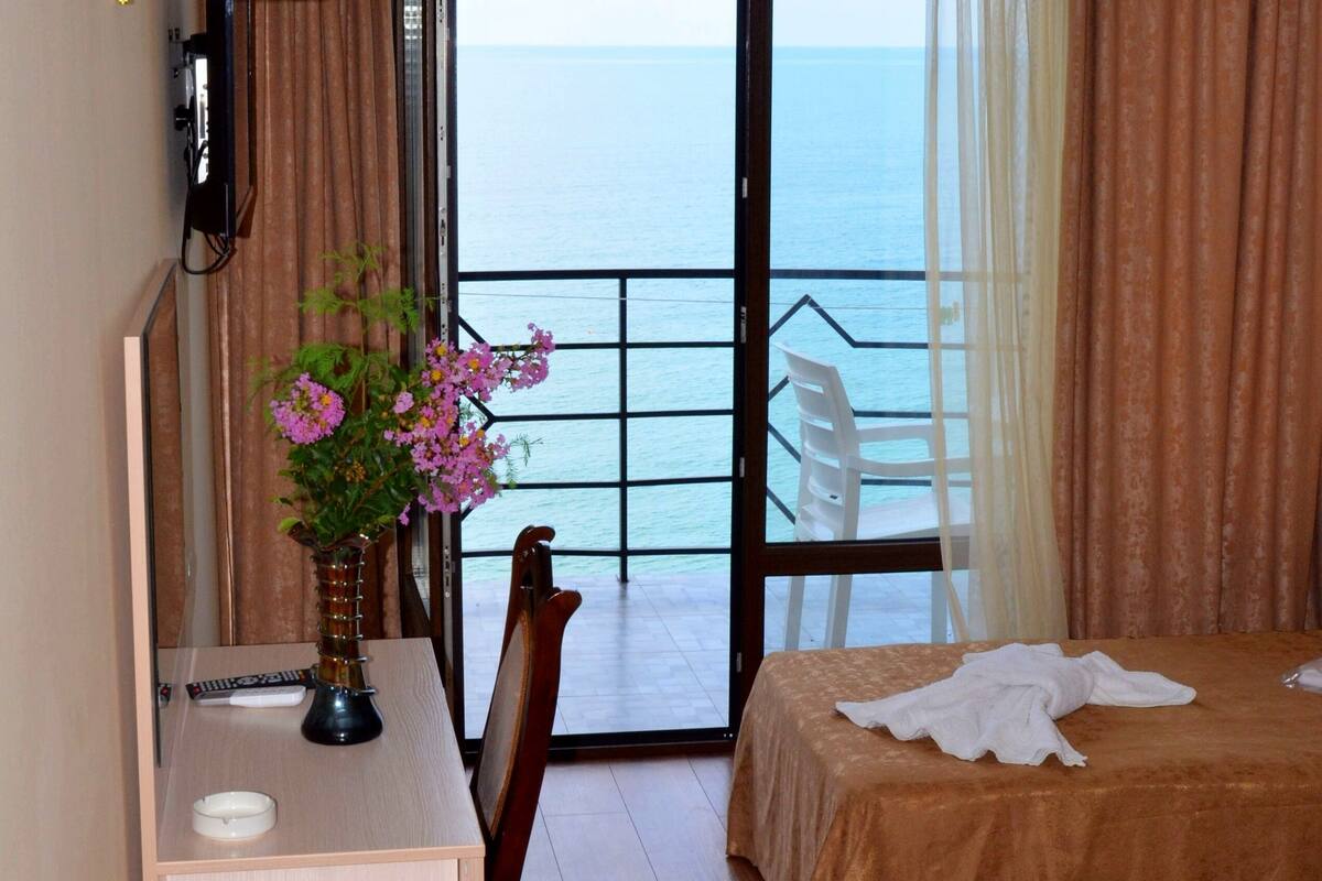 A hotel room with a desk, two beds, a chair, a lamp, and a view of the sea.