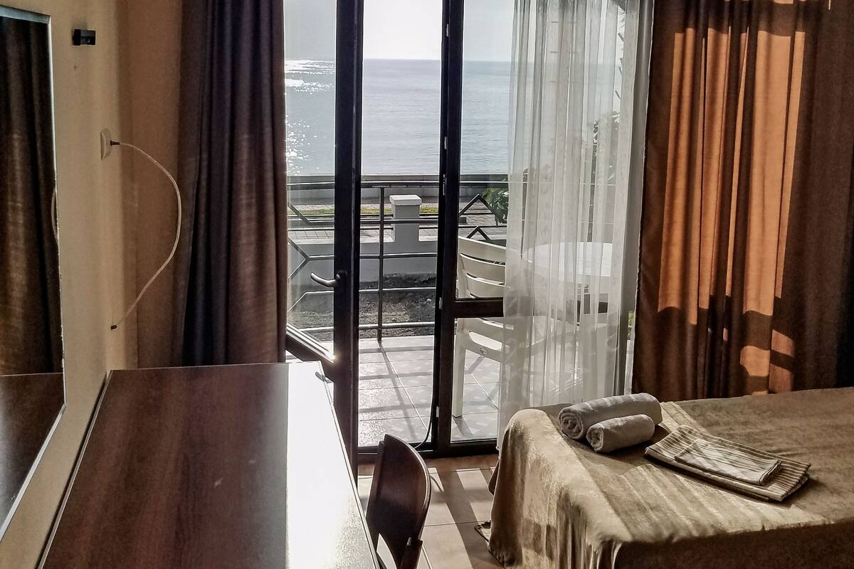 A hotel room with a bed, a desk, and a view of the sea through a large window.