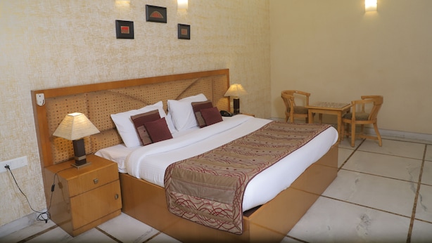A neatly arranged hotel room with a large bed, bedside tables with lamps, a small table and chairs, and a wall with three framed pictures. - Hotel Singh Sahib (New Delhi)
