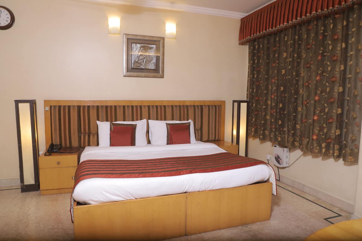 A hotel room with a large bed, a nightstand, a wall clock, a framed picture, and a window with curtains.