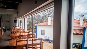 Un espace repas avec des tables et des chaises en bois, de grandes fenêtres et une vue sur un bâtiment dont le toit est recouvert de tuiles rouges.