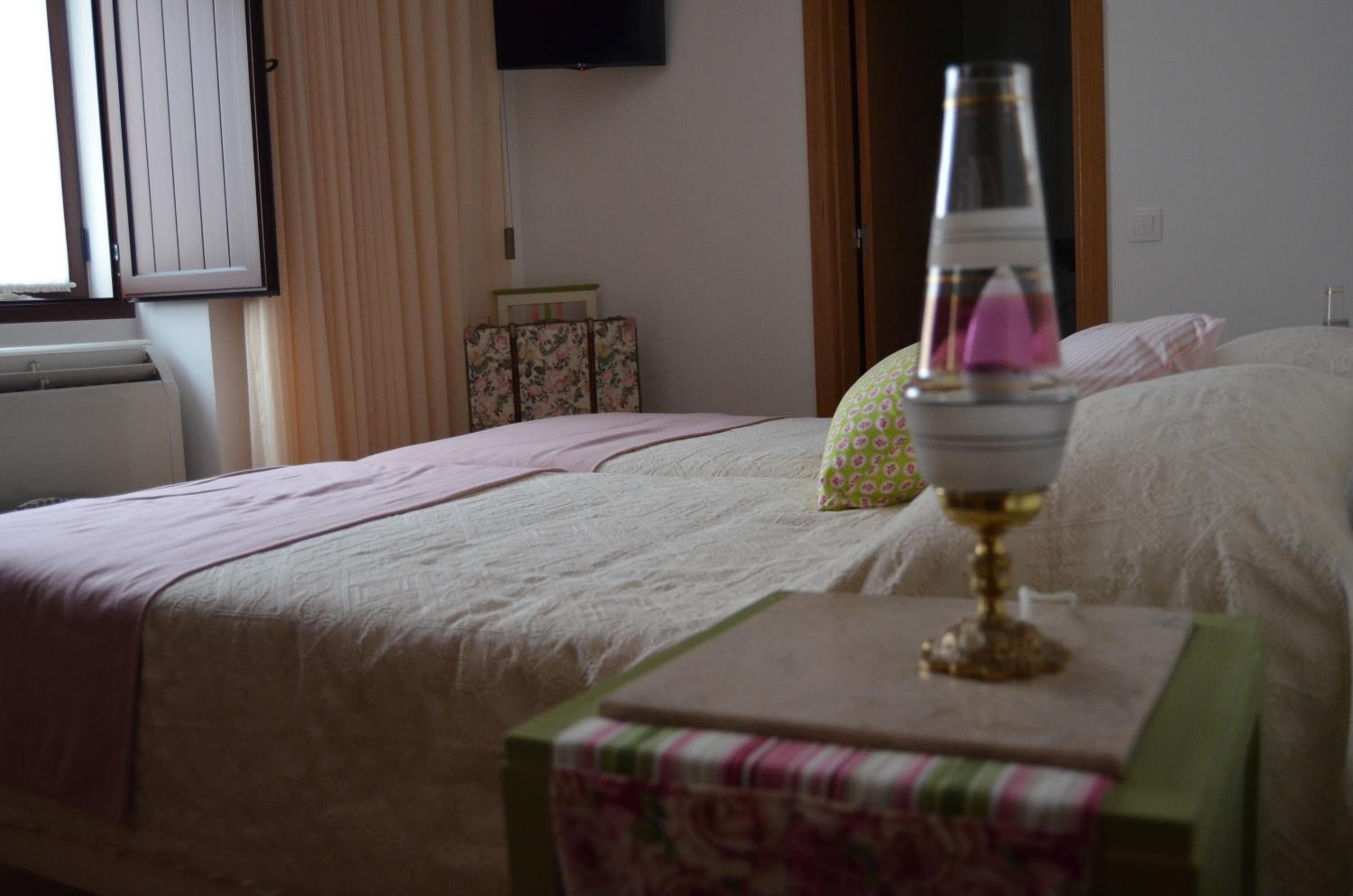 Ein Schlafzimmer mit einem Bett, einem Nachttisch mit Lampe, einem Fenster mit Vorhängen und einem an der Wand befestigten Fernseher.