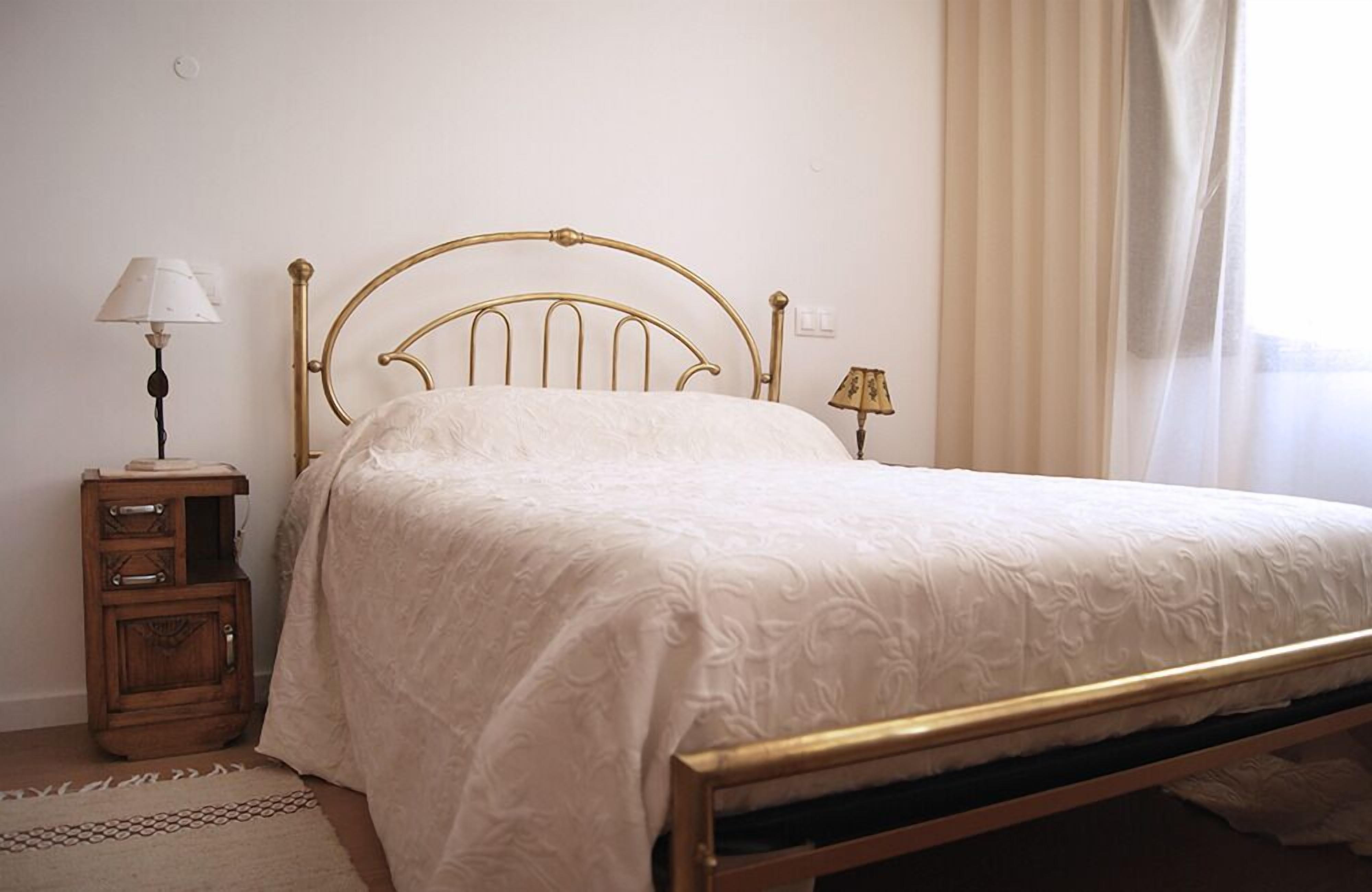 A neatly made bed with a gold frame, a bedside table with a lamp, and a window with curtains.