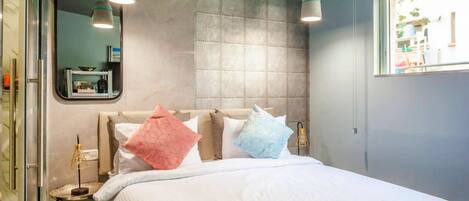 A modern hotel room with a large bed, a bedside table with a lamp, a mirror, and a window with a view of the outside.