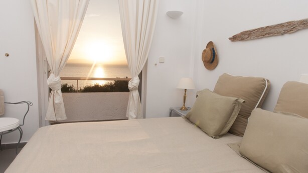 Un dormitorio con cama, cortinas, una mesita de noche, lámpara y vistas al atardecer sobre el mar.