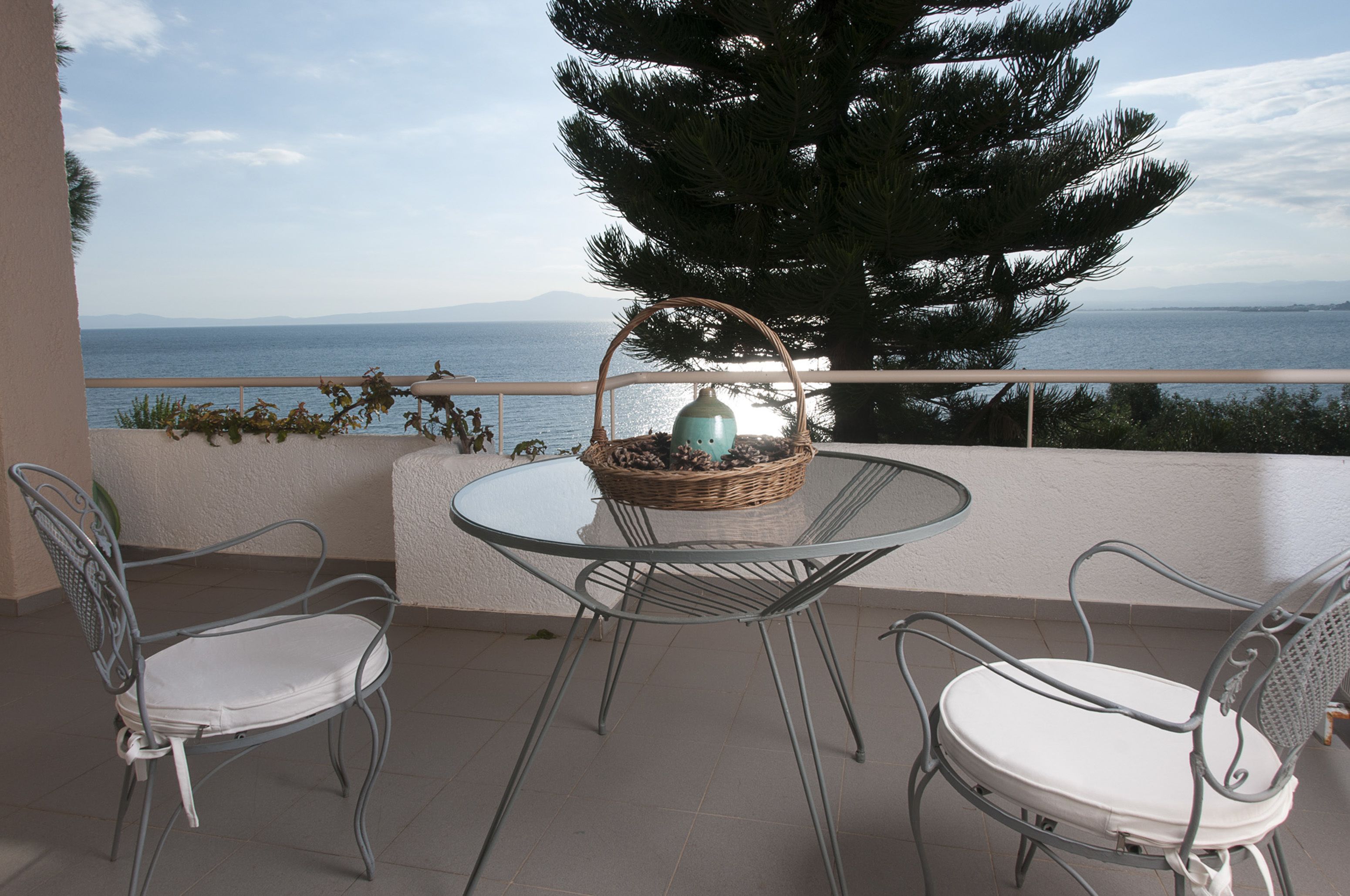 A balcony with two metal chairs and a glass table, a wicker basket with pinecones, and a view of the sea and mountains.