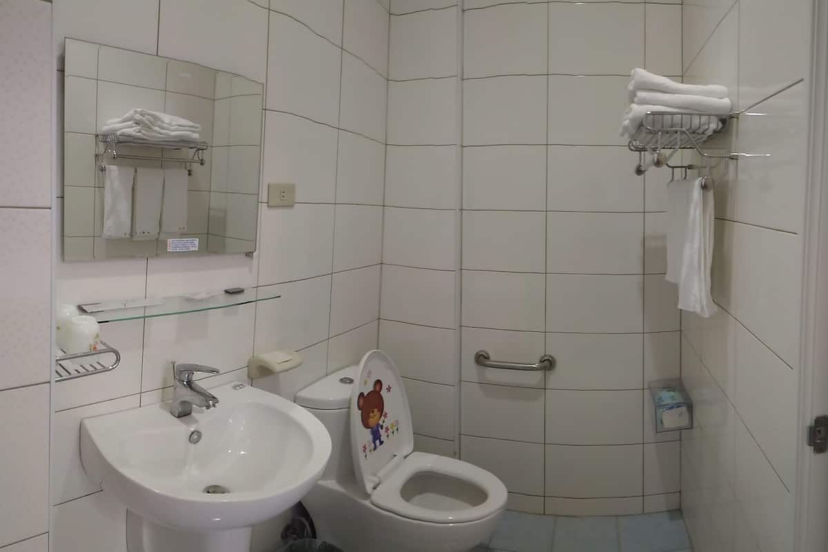 Un baño con lavamanos, inodoro y espejo. Hay toallas en un perchero y un estante de vidrio con artículos de aseo.