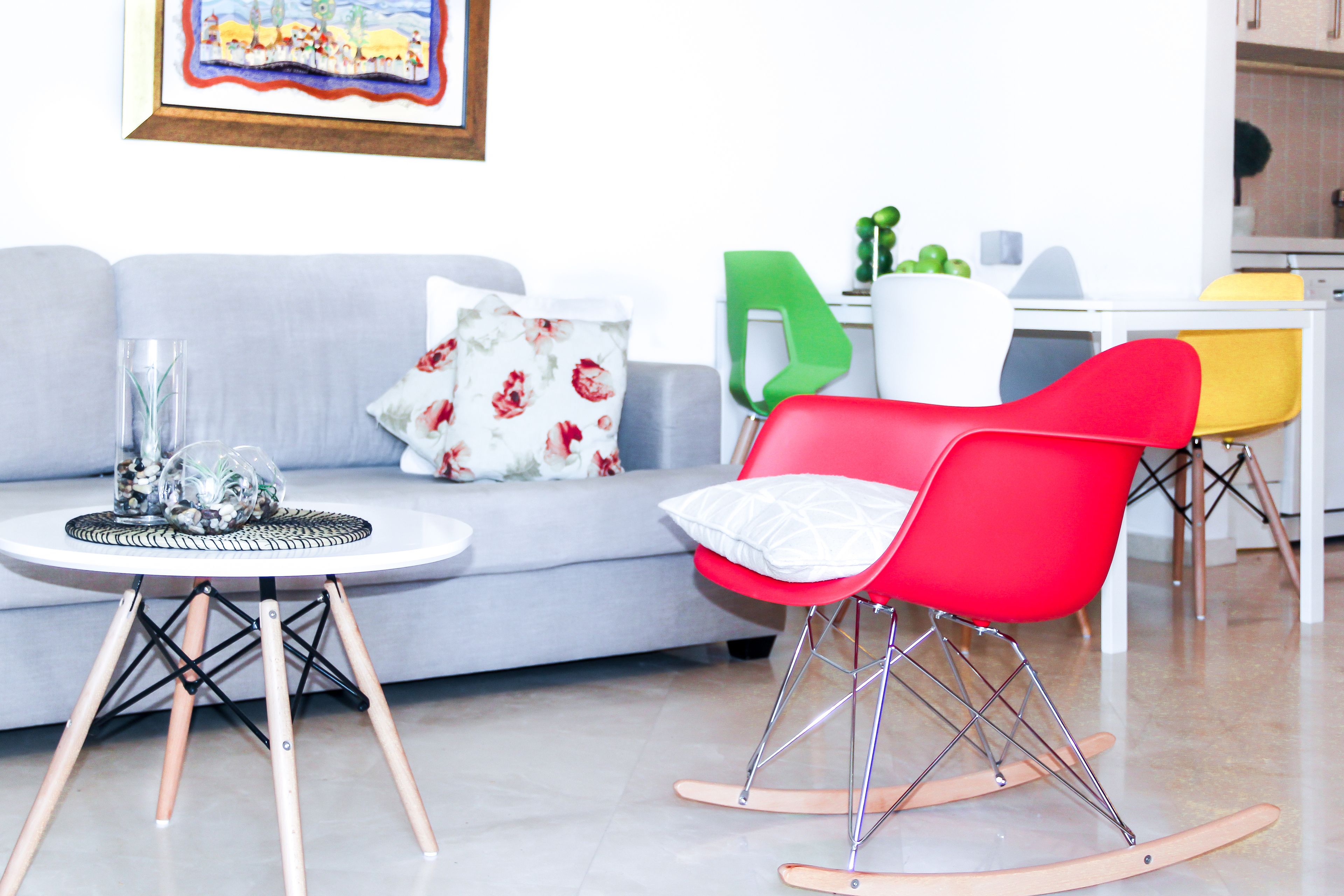 A living room with a grey sofa, a red rocking chair, a small round table with a glass vase, and a green chair.