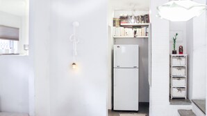 A compact kitchen with a white refrigerator, a small bookshelf, and a hanging light fixture. - Chic Central TLV 1BR Near the Beach (Tel Aviv)