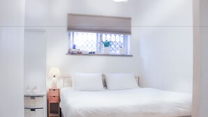 A neatly arranged bedroom with a bed, a bedside table with a lamp, a window with a view, and a shelf with decorative items. - Chic Central TLV 1BR Near the Beach (Tel Aviv)
