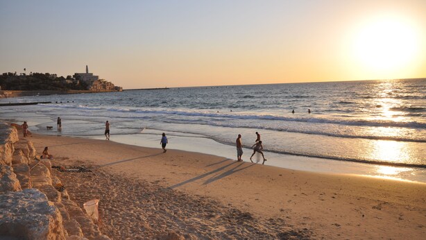 A beach with people walking along the shore, a clear sky, and a distant building on a hill. - Chic Central TLV 1BR Near the Beach (Tel Aviv)