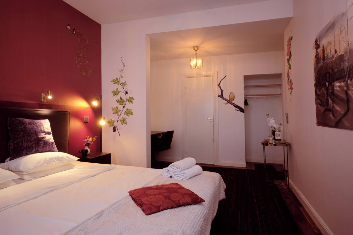 Habitació d'hotel amb un llit gran, una tauleta de nit, una làmpada penjada a la paret, una porta que dóna a un armari i una taula petita amb un vas.