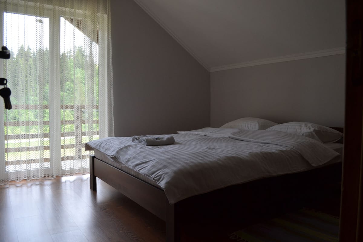 A bedroom with a bed, a window with sheer curtains, and a view of greenery outside.