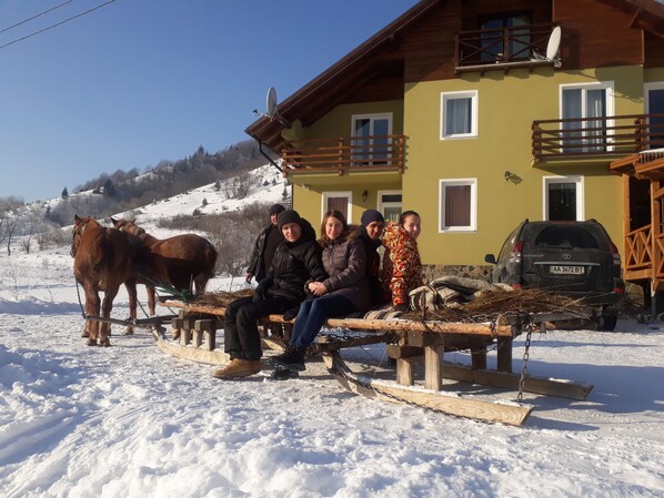 Un groupe de personnes est assis sur un banc devant un bâtiment jaune doté d’un balcon en bois. Un cheval se trouve à proximité, et une voiture est garée à l’arrière-plan.