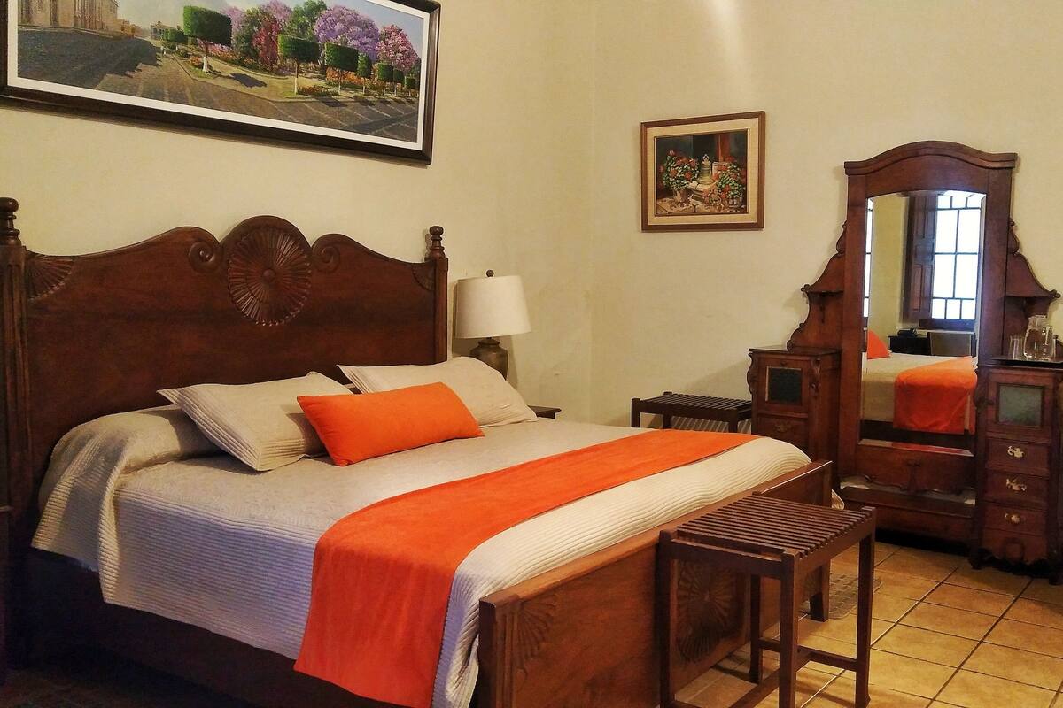 Een keurig opgemaakte slaapkamer met een houten bed, een nachtkastje, een spiegel en een stoel.