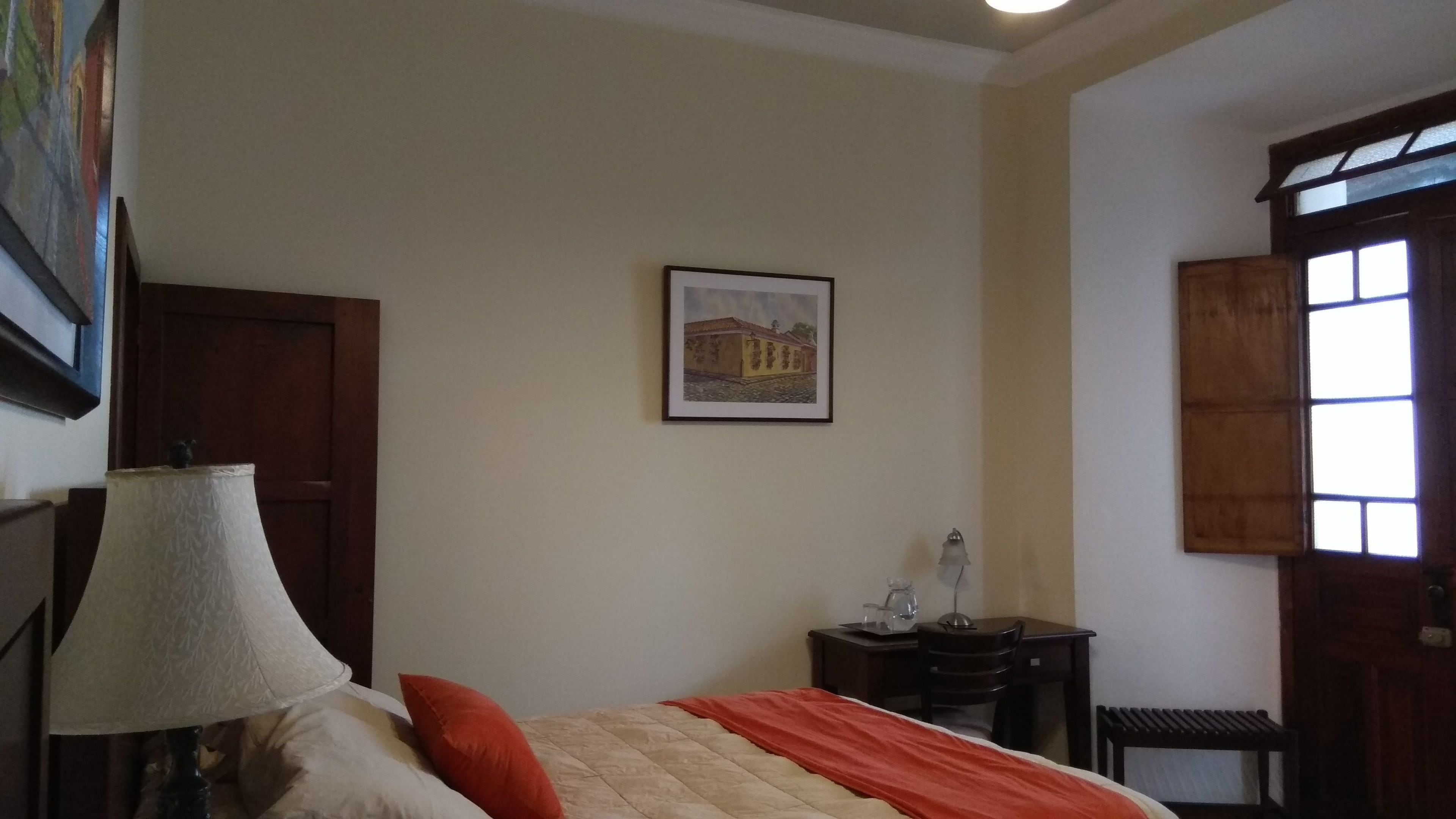 A bedroom with a bed, a lamp, a framed picture on the wall, a desk with a chair, and a window.
