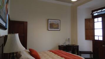 A bedroom with a bed, a lamp, a framed picture on the wall, a desk with a chair, and a window.