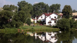 A two-story house with a red roof, surrounded by greenery and a pond. - Pension Všeruby (Vseruby)