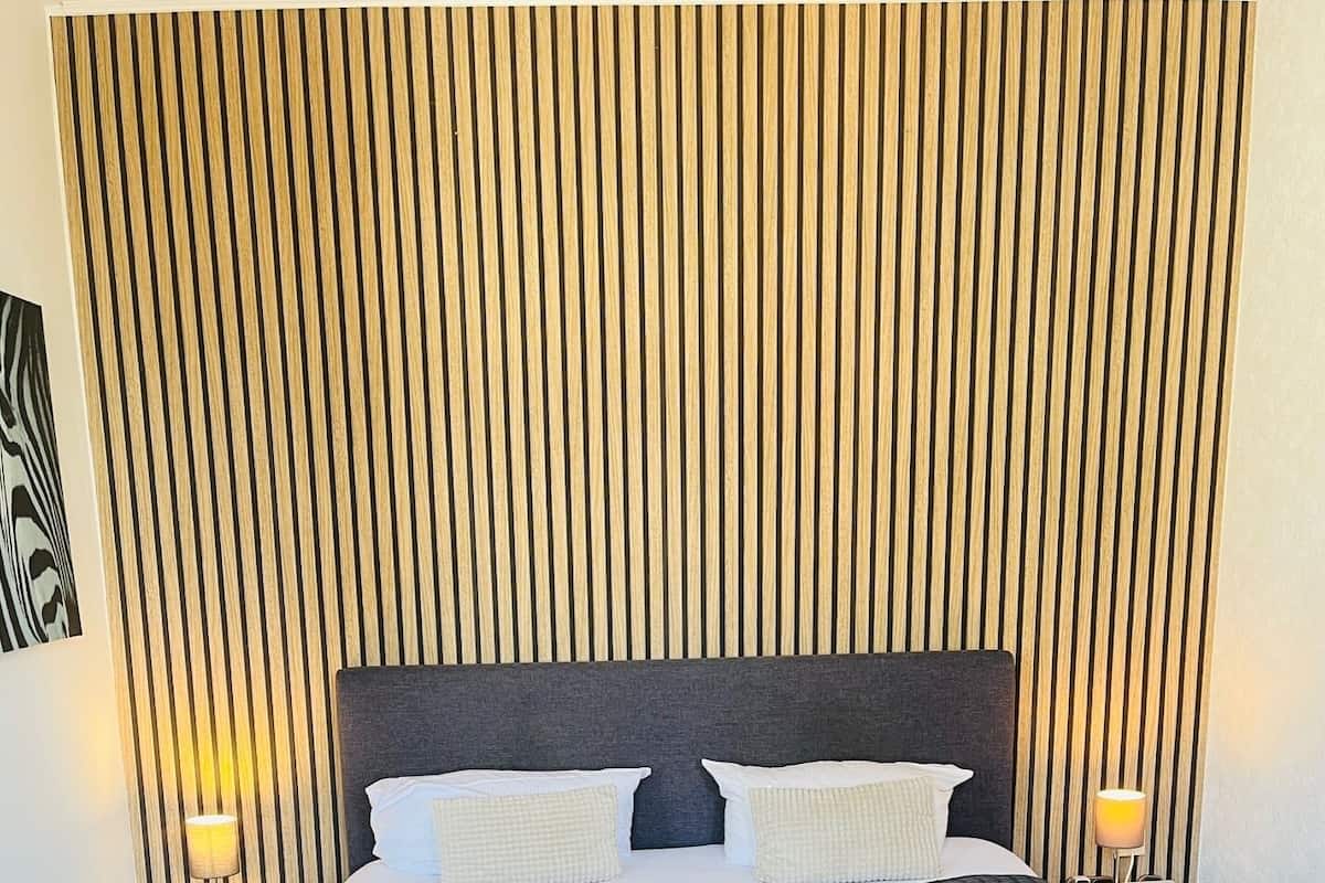 A neatly made bed with a dark blue quilt, two white pillows, and two folded towels on the bed. There are two bedside tables with lamps, and a wooden slat wall behind the bed.