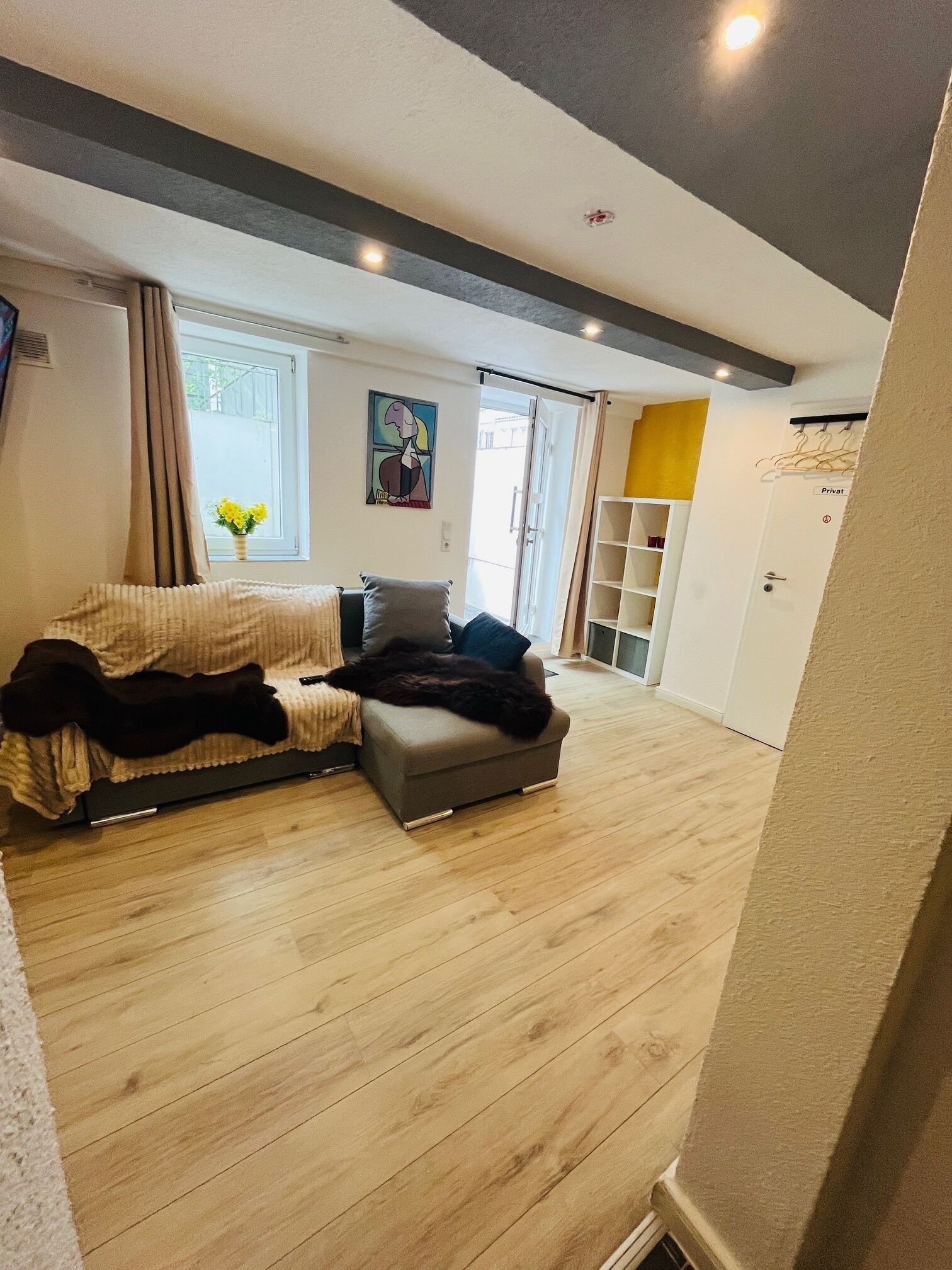 A modern living room with a grey sofa, a small ottoman, and a wooden floor. There is a window with curtains, a painting on the wall, and a door labeled "Privat".