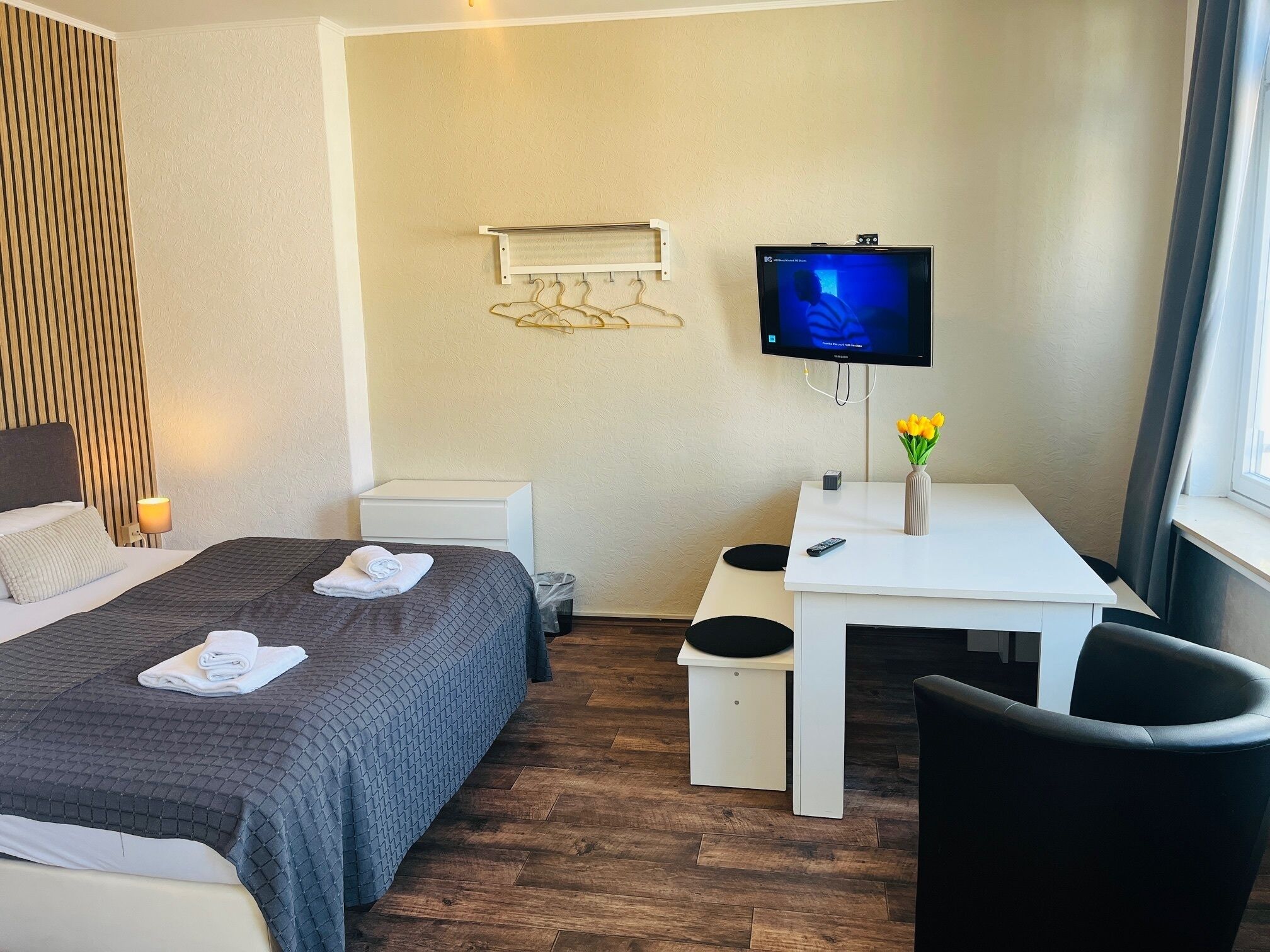 Hotellihuone, jossa on sänky, työpöytä, tuoli, televisio ja pieni pöytä, jonka päällä on kukkia.