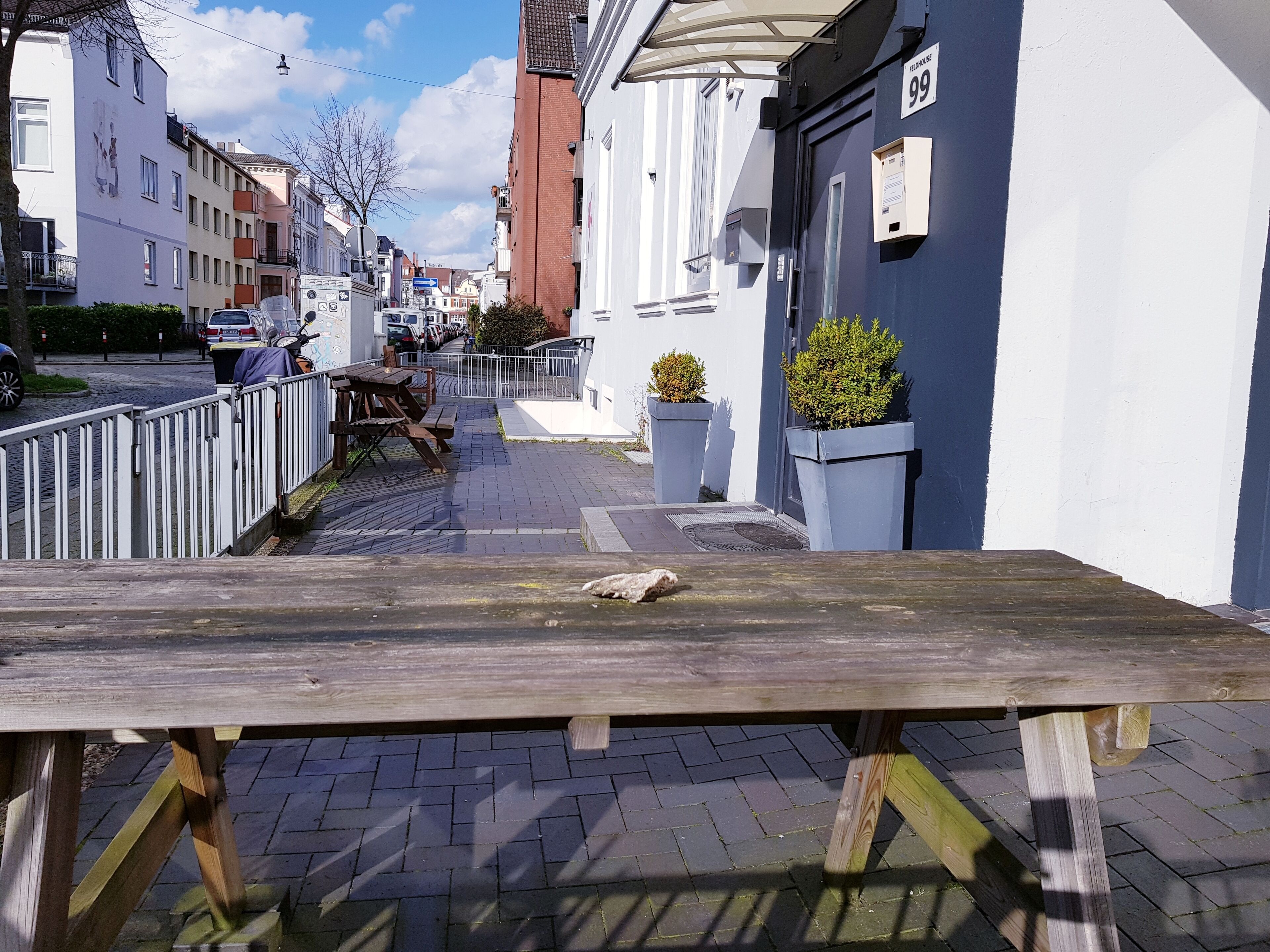 Une rue étroite, avec un banc en bois au premier plan, une rangée de voitures garées et des immeubles dotés de balcons et d'enseignes.